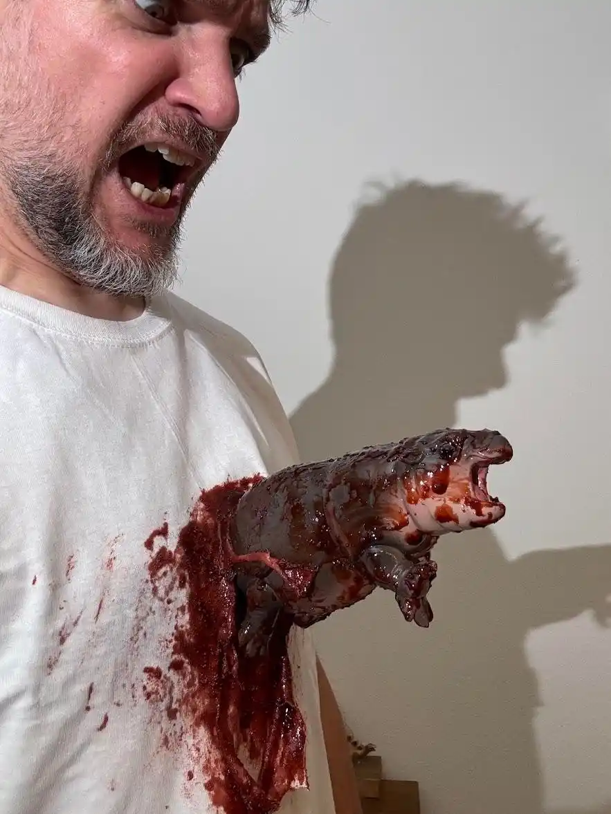 A man with a beard and white t-shirt is screaming in shock as a bloody, grotesque salamander appears to be bursting out of…