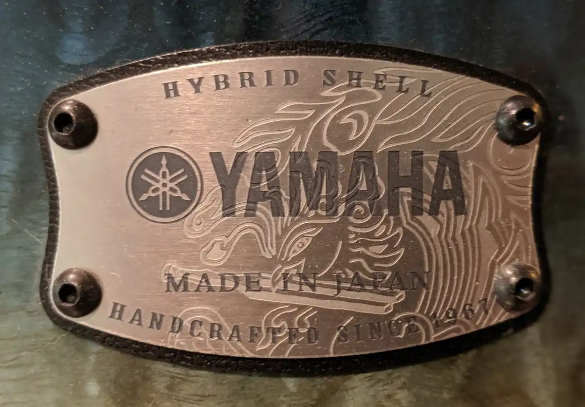 A close-up of a Yamaha drum shell badge showing 'HYBRID SHELL', the Yamaha logo, 'MADE IN JAPAN', and 'HANDCRAFTED SINCE…