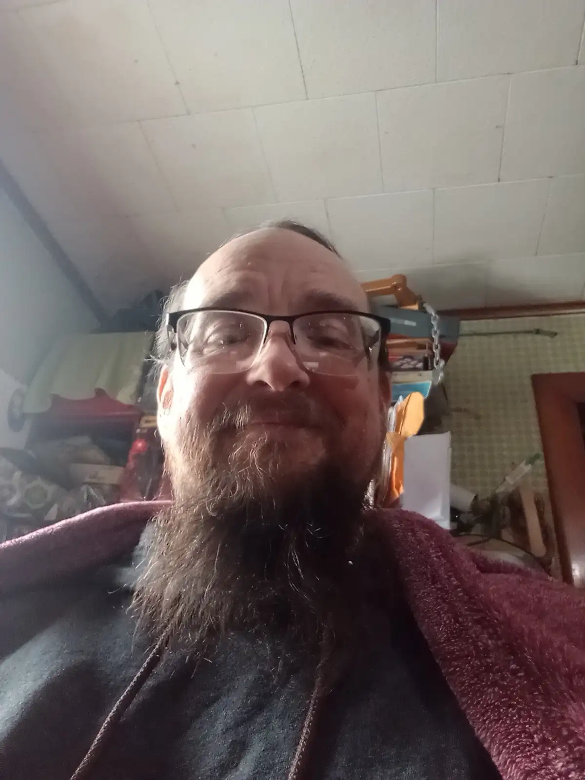 A man with glasses and a long beard smiles at the camera while wearing a dark hoodie and a maroon blanket draped over his…