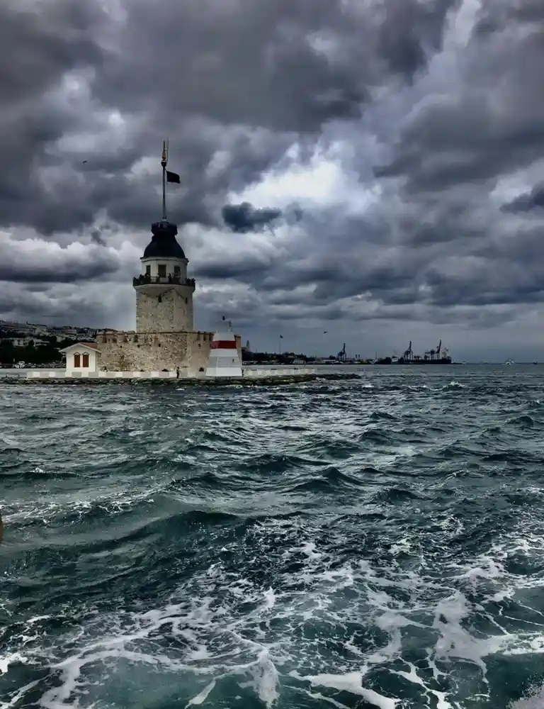 A stormy seascape featuring the historic Maiden's Tower in Istanbul, with dark, turbulent waters in the foreground and a…