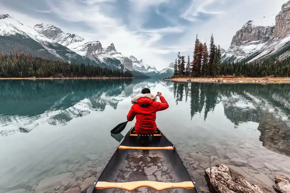A person in a red winter jacket sits in a canoe on a calm, clear lake, holding a phone to take a photo. Snow-capped…