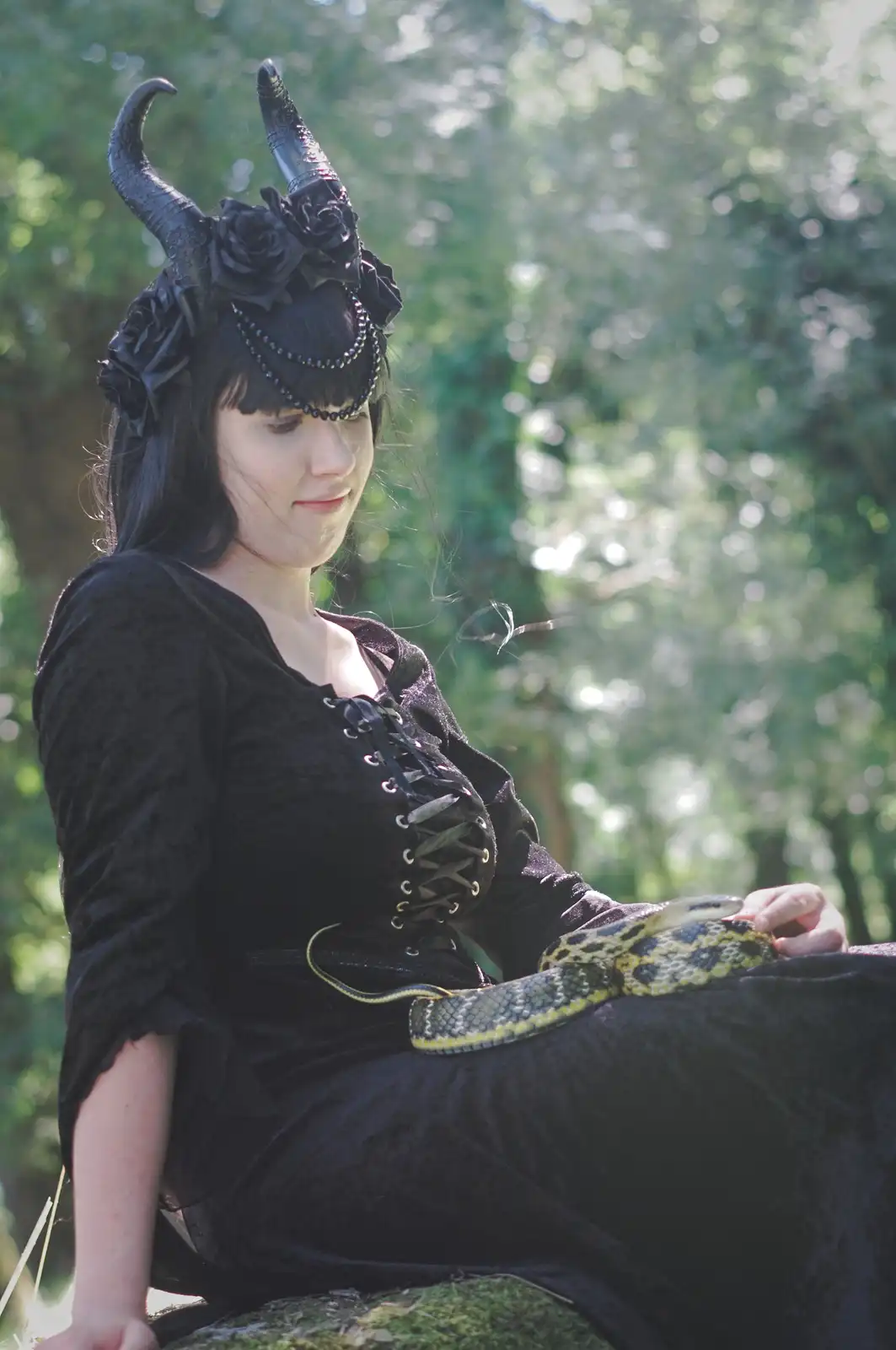 A woman dressed in black gothic attire with horns and roses on her head sits outdoors holding a snake. The overlaid text…