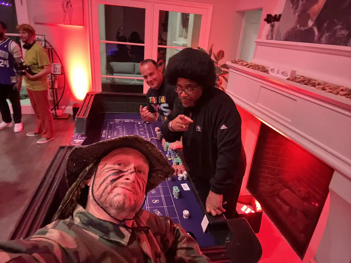 A man in camouflage clothing and a tiger-striped face paint takes a selfie at a casino table, while two other men stand…