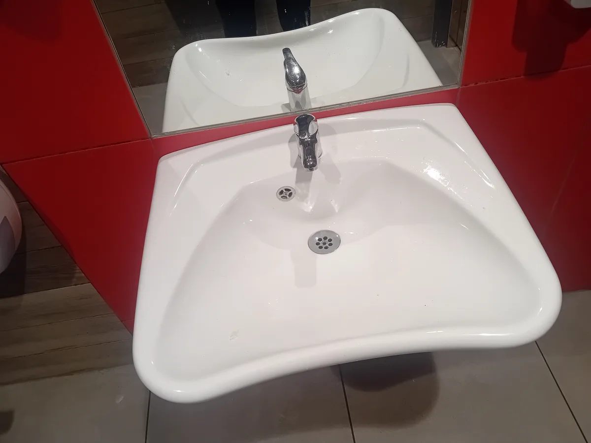 A white, oddly shaped wall-mounted sink with a chrome faucet and drain, set against red walls and a mirror above, with the…
