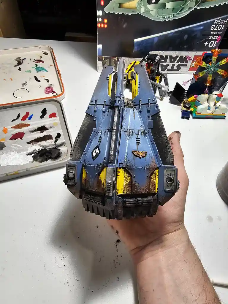 A hand holds a heavily weathered, blue and yellow painted Warhammer 40k-style model spaceship with dirt and chipped paint…