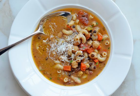 a top-down photo of a white bowl filled with a hearty-looking pasta soup containing elbow macaroni, diced carrots, black…