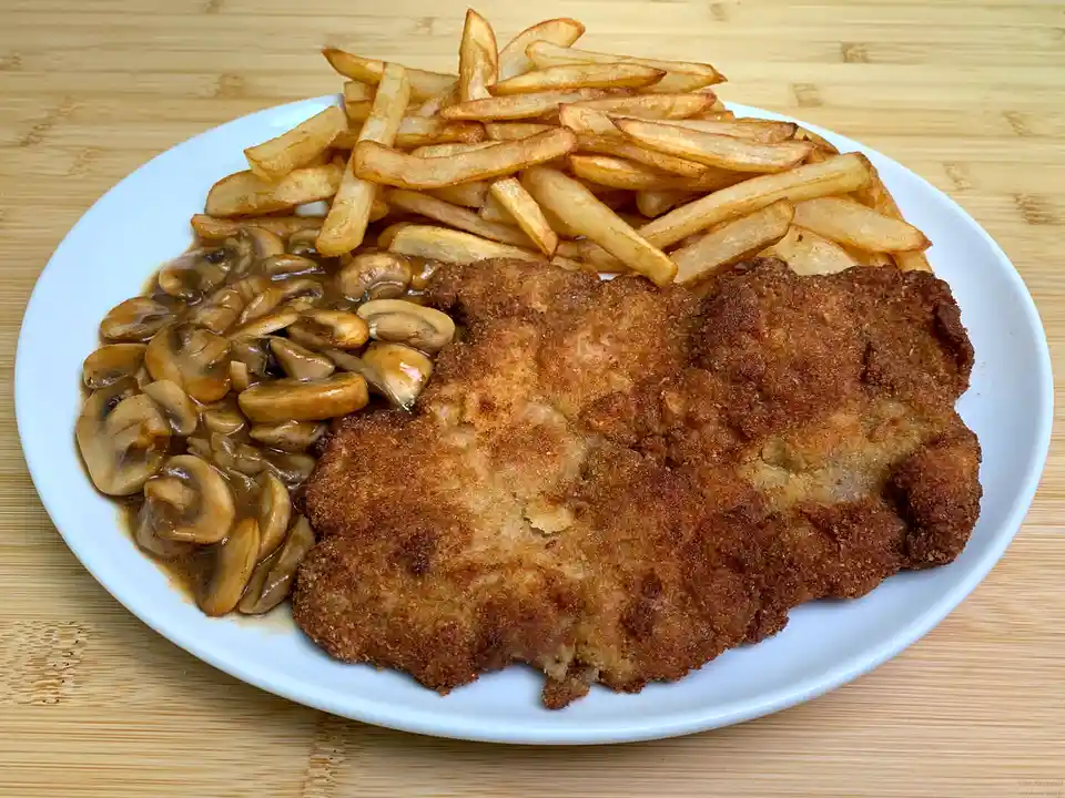 A plate of food featuring a large breaded and fried cutlet, a serving of french fries, and a side of mushrooms in gravy, all…