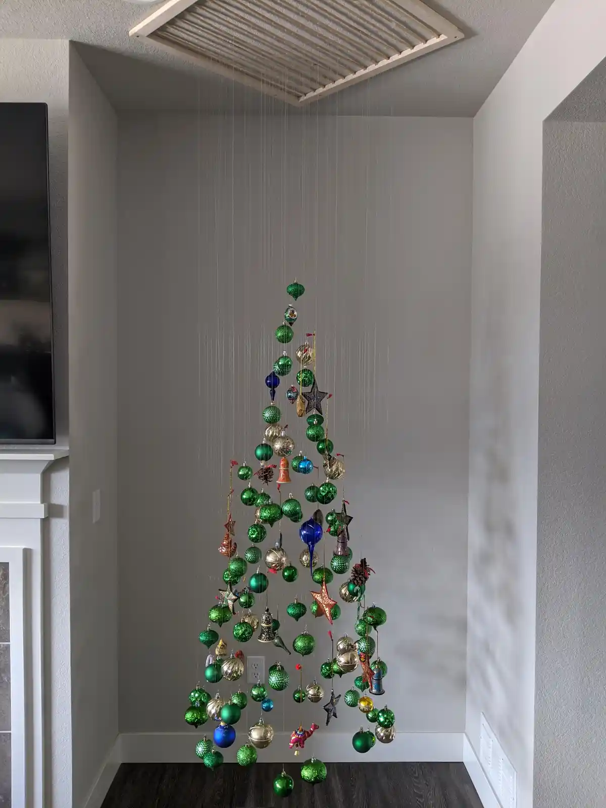 A Christmas tree made entirely of hanging ornaments suspended from a ceiling vent in a corner of a room, with a TV and…