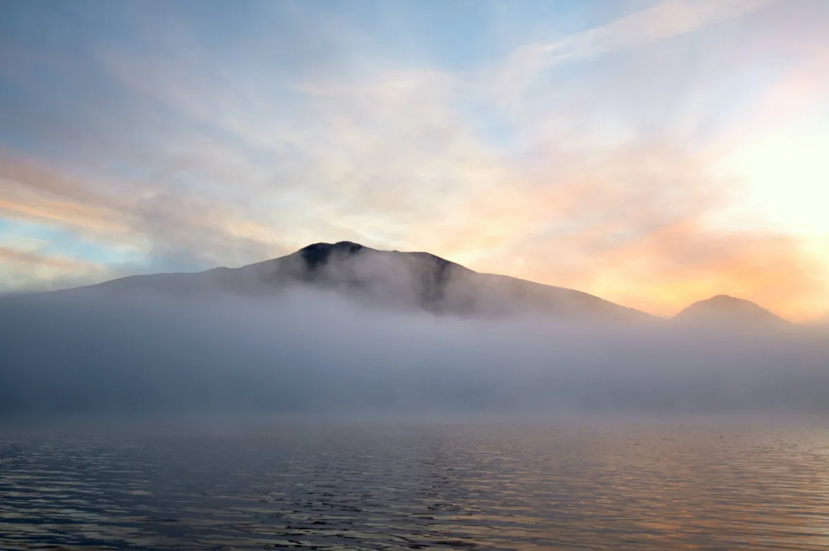 A serene landscape photo shows a mountain partially obscured by fog over a calm body of water, with a sunrise or sunset…