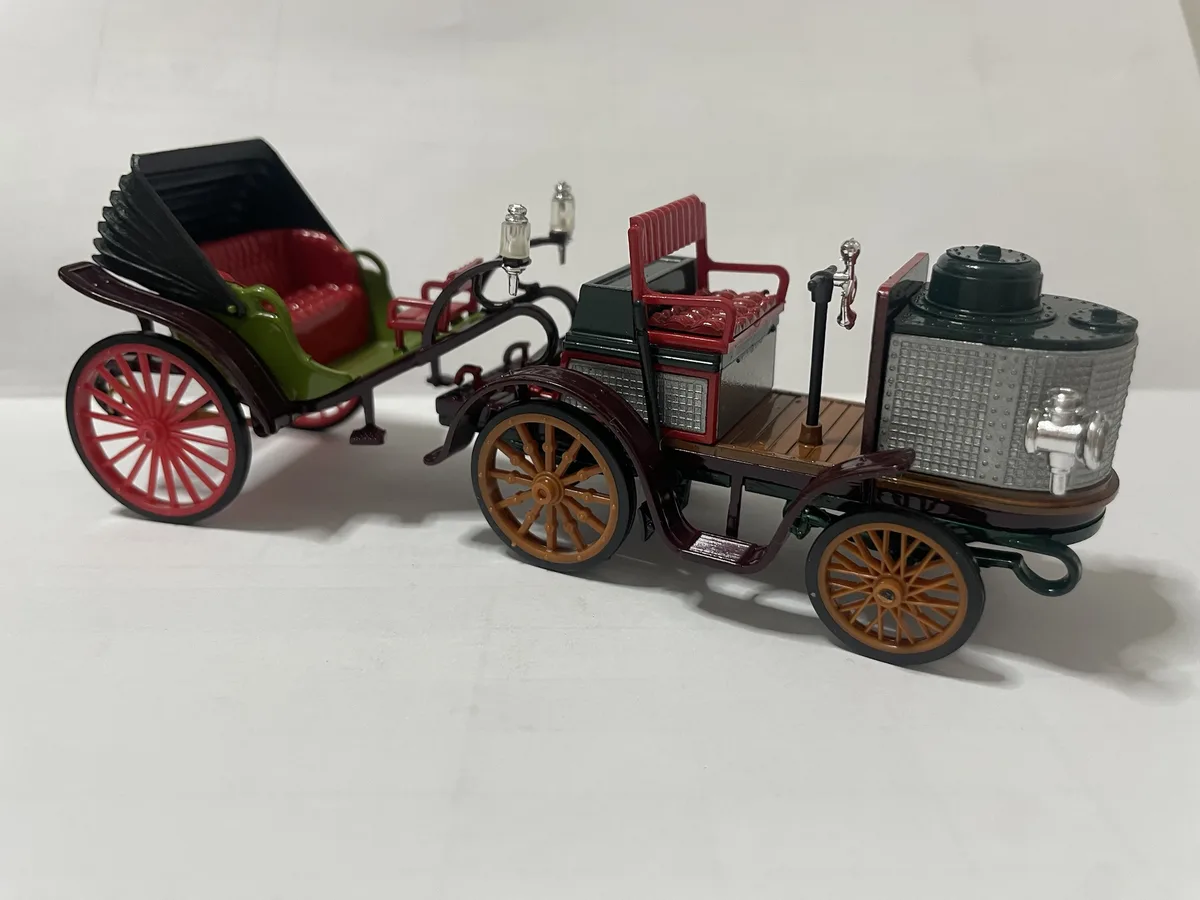 A toy model of a vintage steam-powered carriage with a passenger compartment and a large boiler engine at the rear, set…