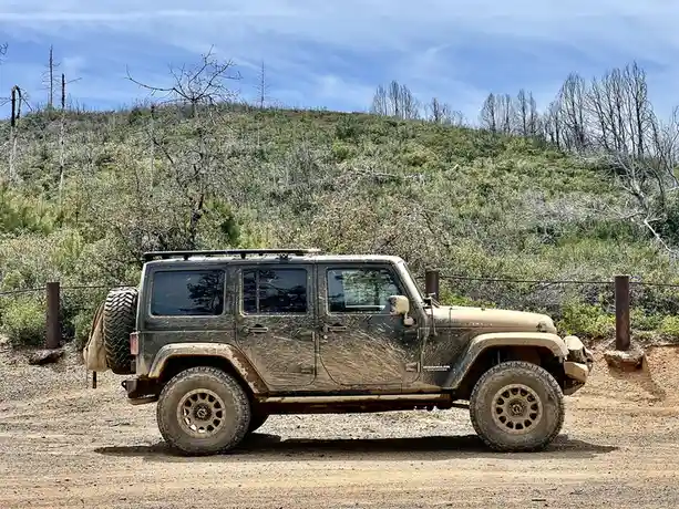 A heavily mud-splattered dark green Jeep Wrangler Unlimited is parked on a dirt road in a rugged, brush-covered landscape…