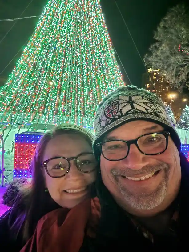 A selfie of a smiling man and woman in front of a large, brightly lit Christmas tree at night. The man wears a patterned…