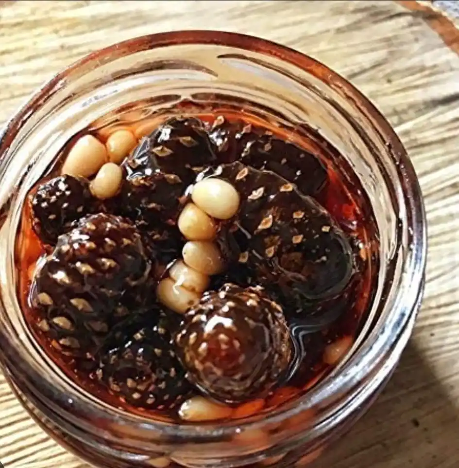 A close-up photo shows a clear glass jar filled with dark, glossy figs submerged in a thick, dark syrup, mixed with…