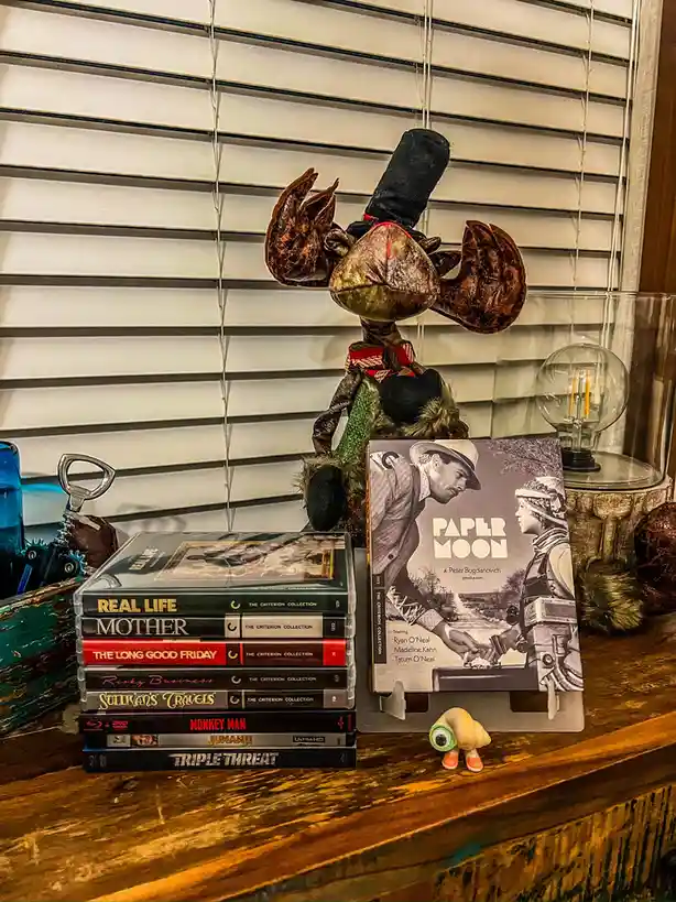 A stack of Criterion Collection movie cases sits on a wooden shelf next to a large plush moose wearing a top hat and bow…