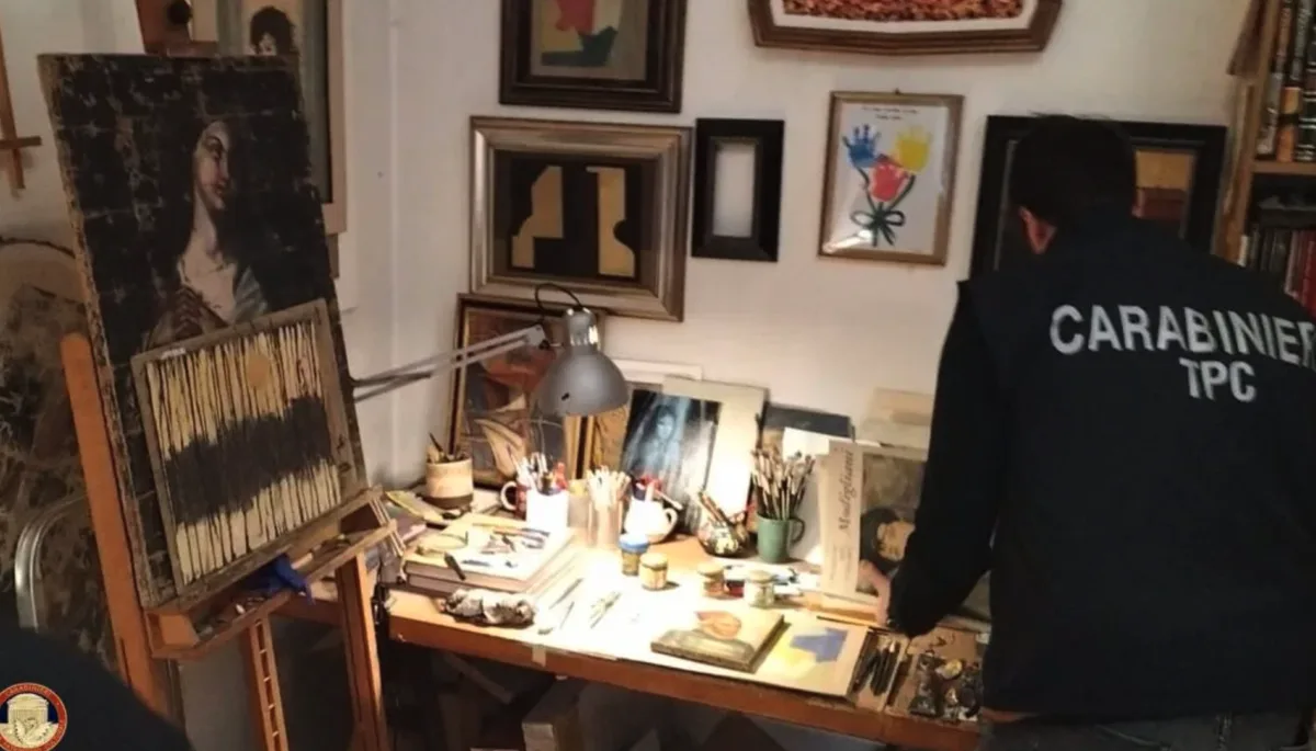 A person wearing a dark blue jacket with 'CARABINIER TPC' printed on the back stands in an artist's cluttered studio, facing…