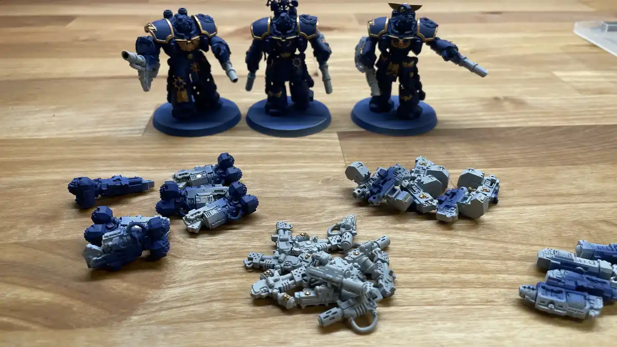 Three painted blue and gold space marine miniatures stand upright on a wooden floor, while numerous unpainted grey weapon…