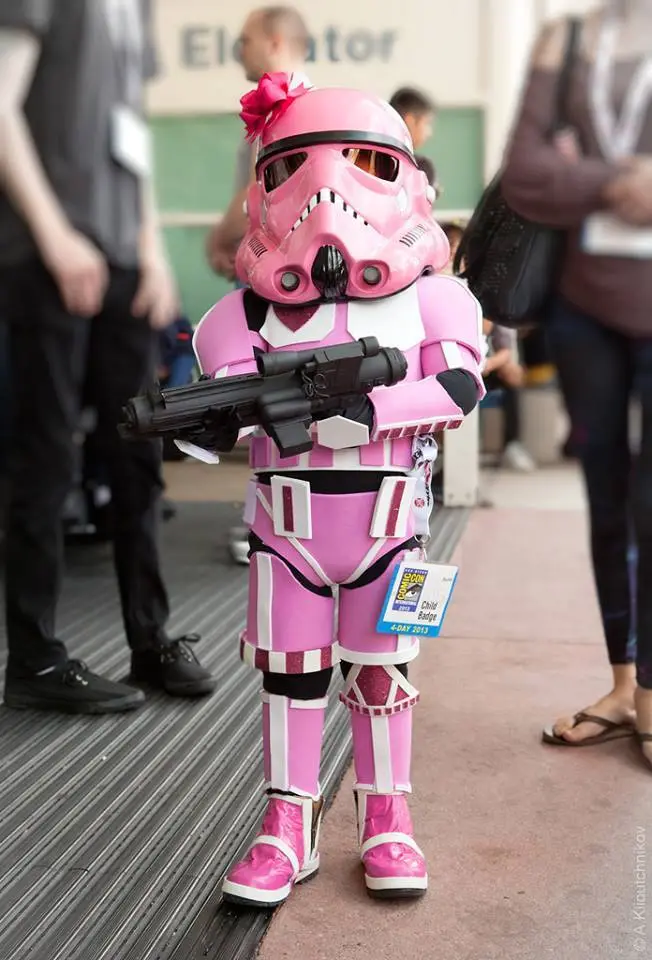 A child in a full pink stormtrooper costume from Star Wars stands holding a blaster, with a large pink bow on their helmet…