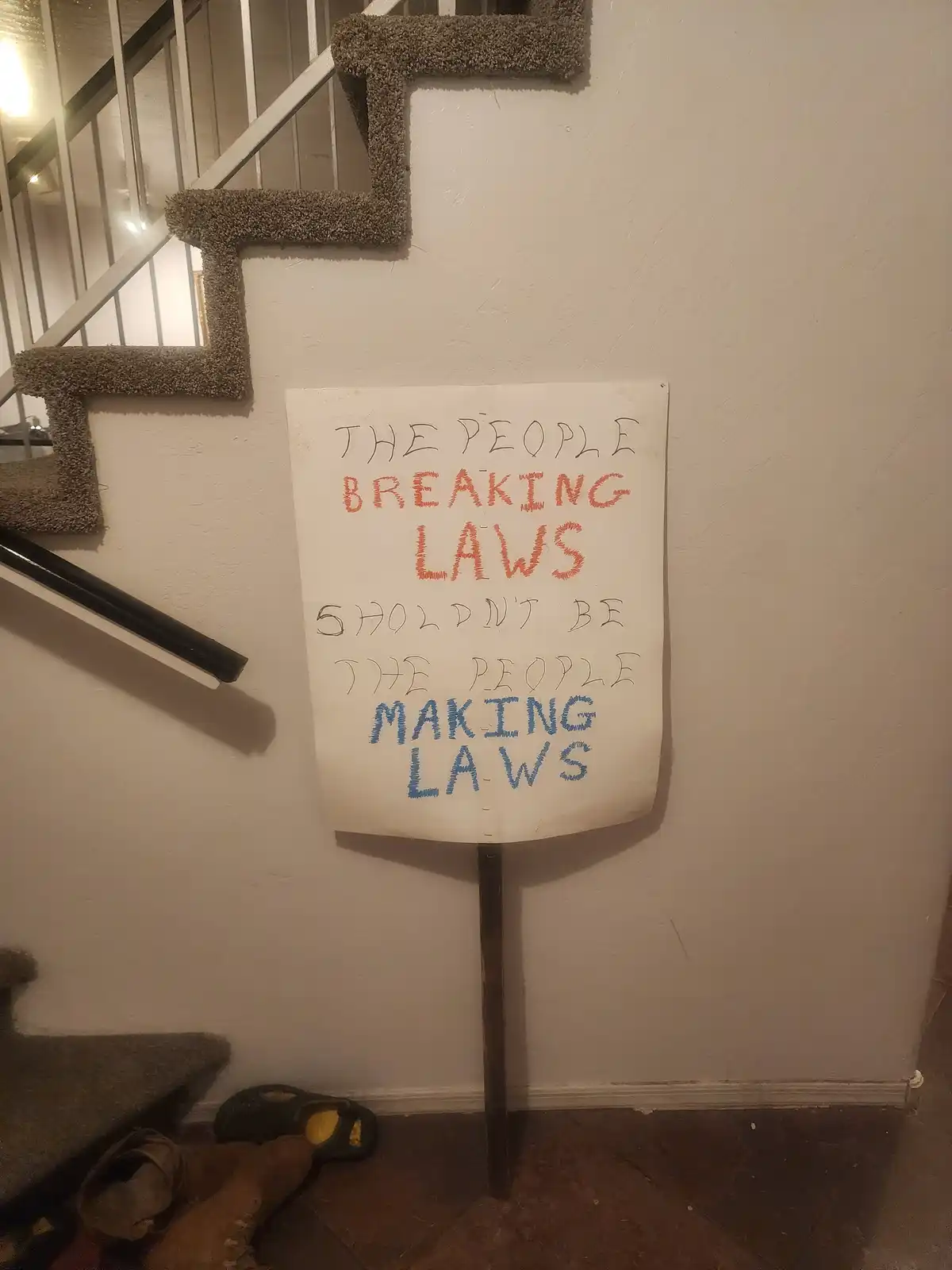 A handmade sign with handwritten text in black, orange, and blue marker is propped against a wall next to a carpeted…
