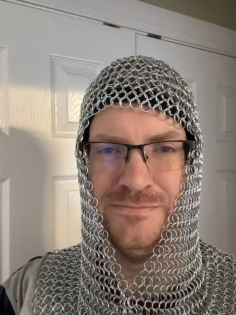 A man with glasses and a mustache wears a full chainmail coif and gorget over his head and shoulders, standing in front of a…