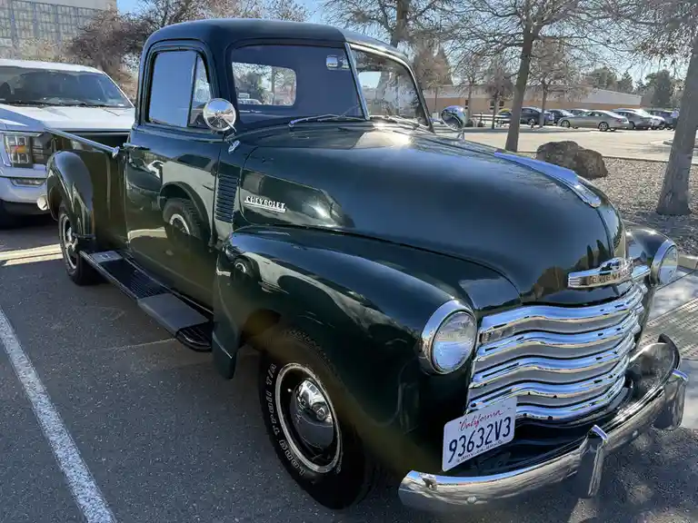 A shiny, dark green vintage Chevrolet pickup truck is parked in a lot, its chrome grille and bumpers gleaming under…