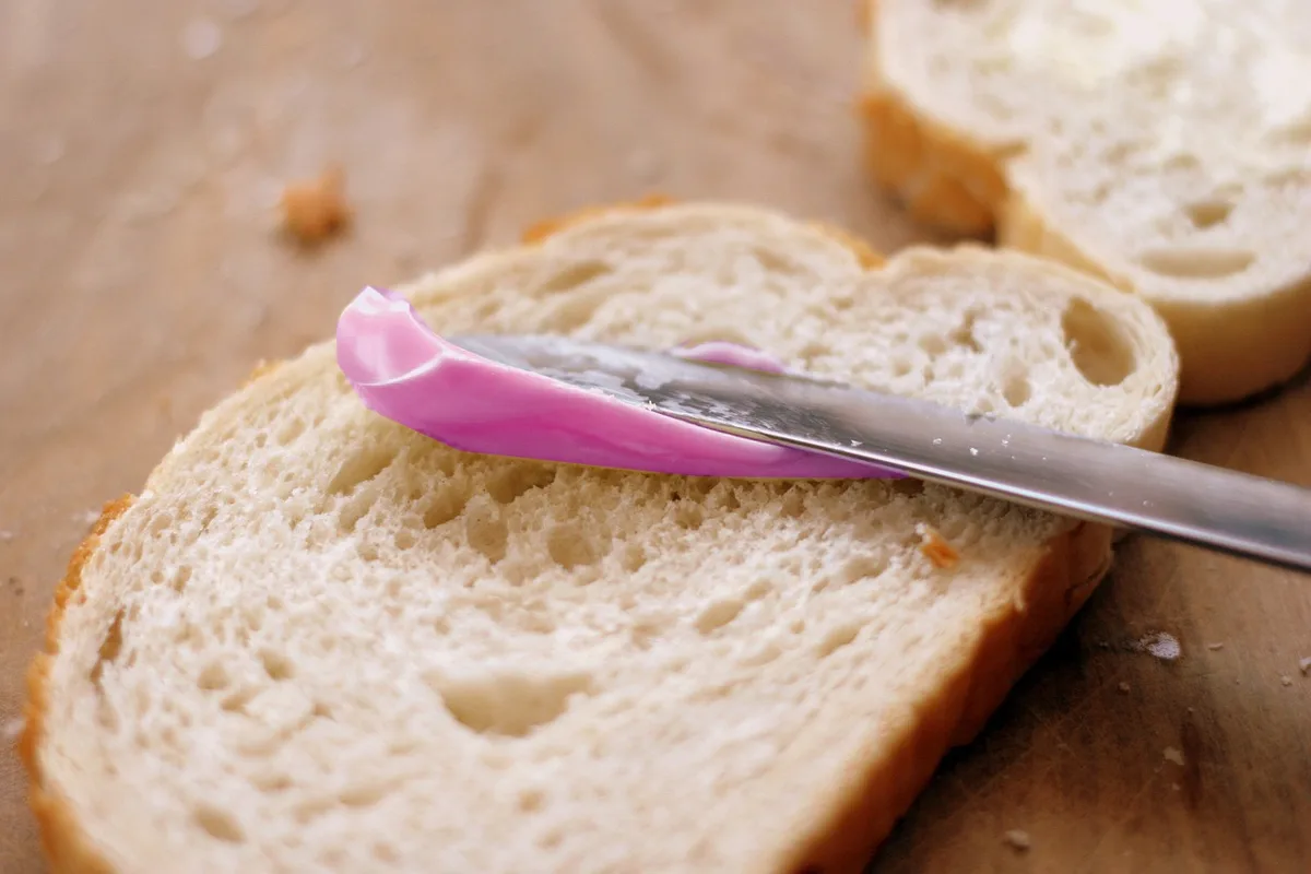 A close-up photo shows a butter knife with a pink plastic handle resting on a slice of white bread, with another slice…