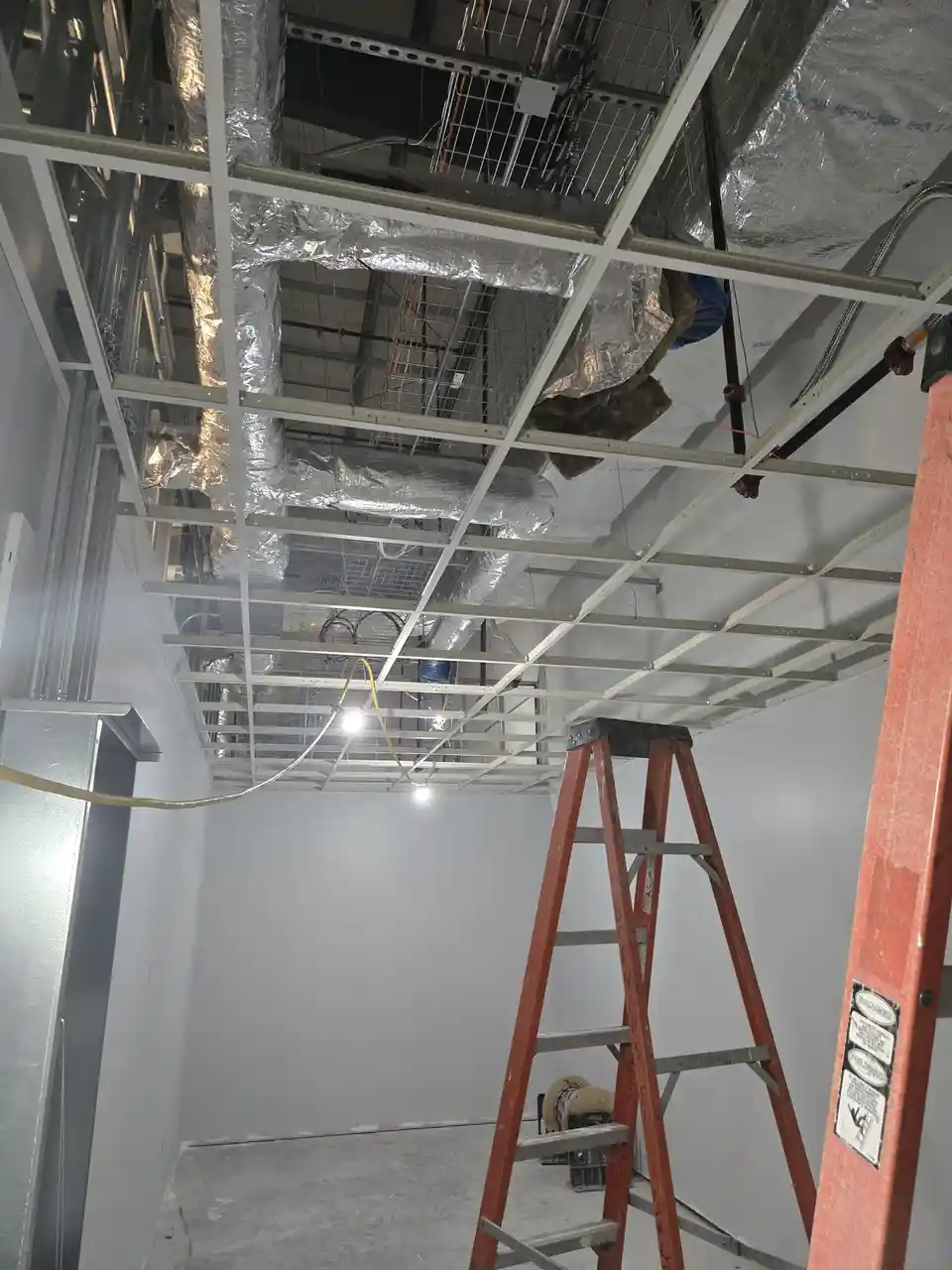 A photo of a room under construction with exposed ceiling framing, ductwork, and wiring, featuring a red ladder in the…