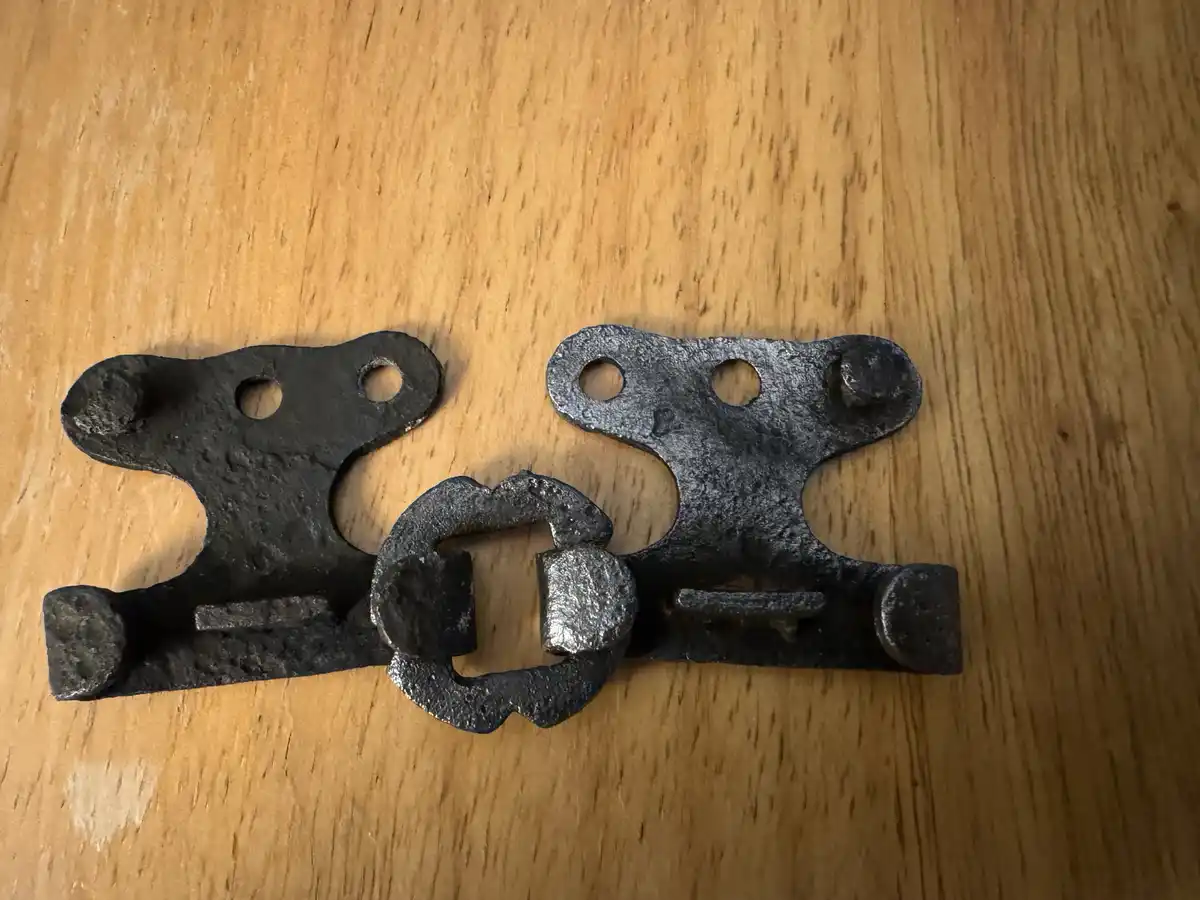 A close-up photo shows a rustic, dark metal latch mechanism with two hinged pieces and a central locking bar, resting on a…