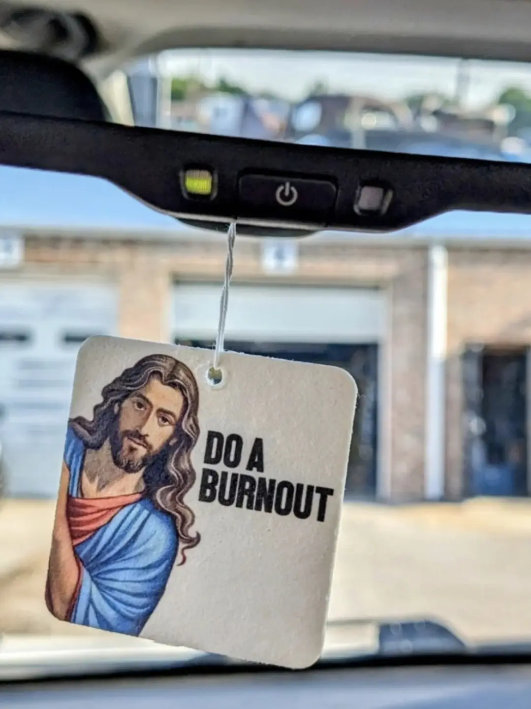 A square car air freshener hangs from a rearview mirror, featuring a depiction of Jesus Christ in a blue and red robe, with…