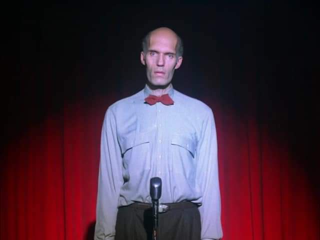 A bald man in a light blue shirt and red bow tie stands at a microphone on a stage with red curtains behind him, looking…