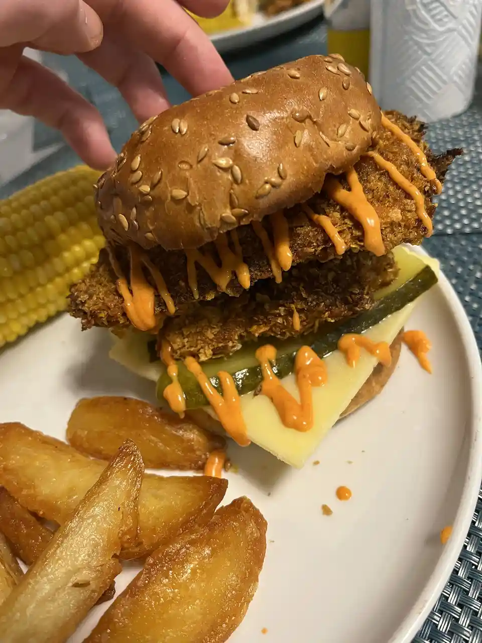 A close-up photo shows a hand holding a large, messy double fried chicken sandwich on a sesame seed bun, drizzled with…