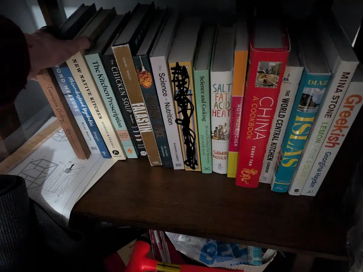 A hand reaches toward a bookshelf filled with numerous cookbooks, including titles like 'Salt Fat Acid Heat,' 'China A…