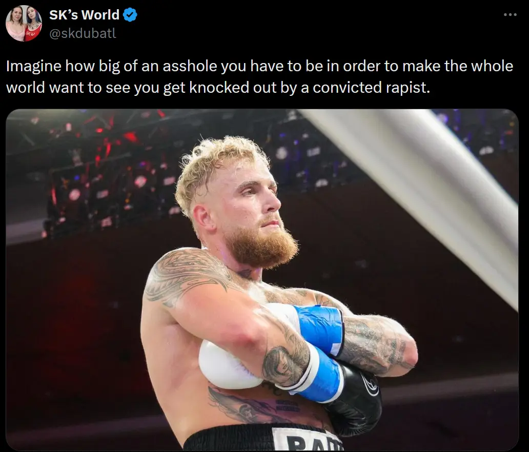 A shirtless, tattooed male boxer with a beard and blonde hair stands in a boxing ring, arms crossed over his chest, wearing…