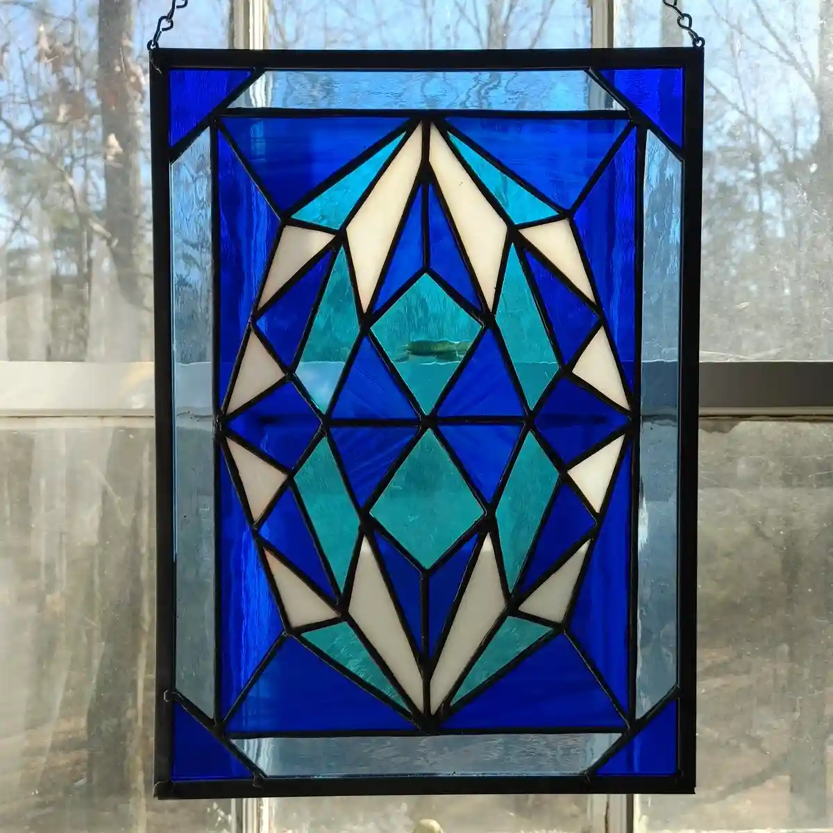 A rectangular stained glass window with a geometric diamond pattern made of blue, teal, and cream-colored glass pieces…