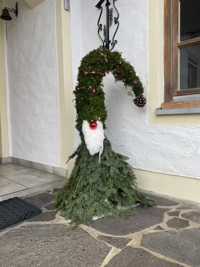 A festive outdoor decoration shaped like a snowman, constructed from green pine branches, features a white fluffy beard made…