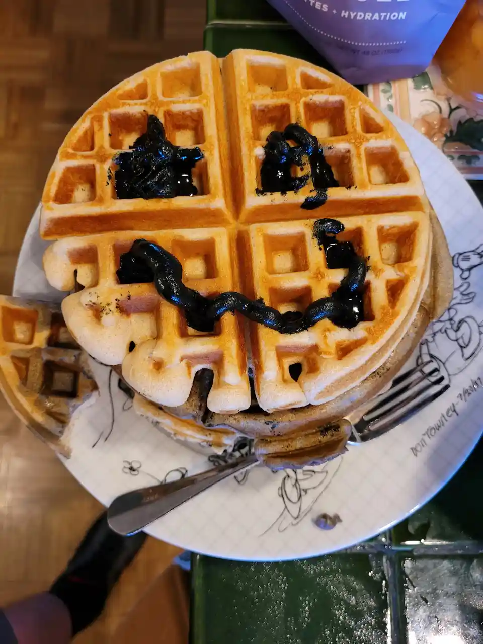 A golden-brown waffle on a white plate is topped with dark, glossy syrup arranged to look like a smiling face with two eyes…