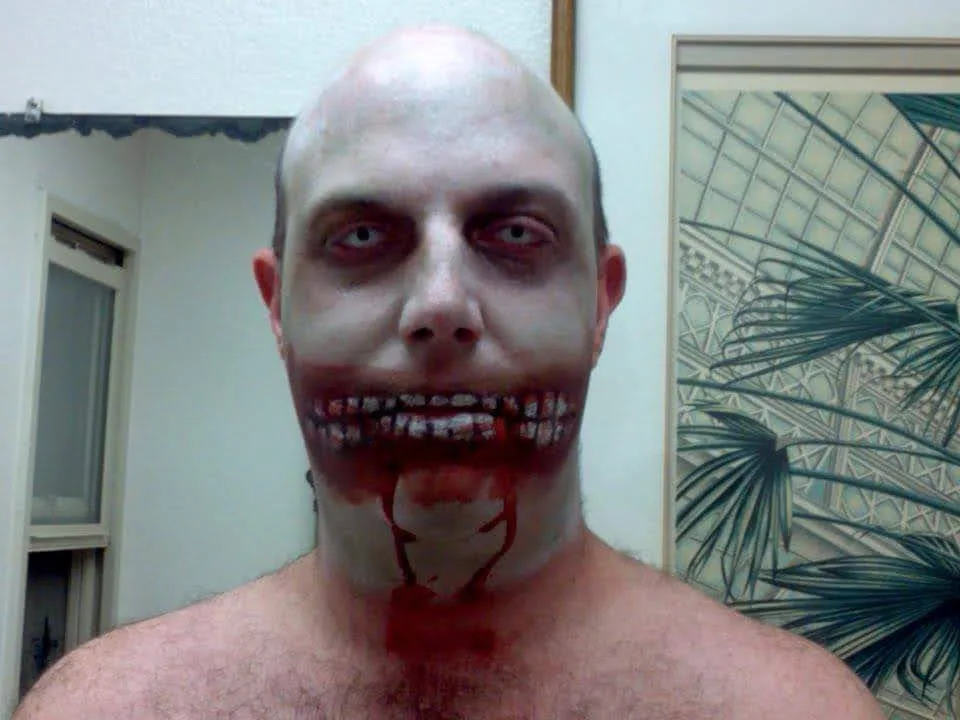 A man with a bald head and pale makeup stares directly at the camera, his mouth grotesquely altered to appear as if he has…