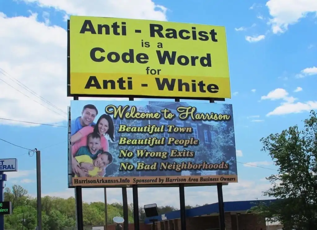 A large yellow billboard reads 'Anti - Racist is a Code Word for Anti - White' above a 'Welcome to Harrison' sign featuring…