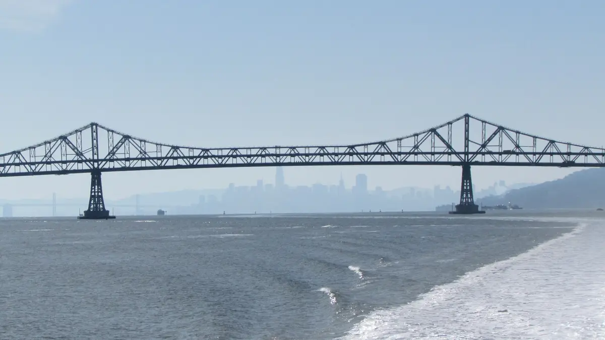 A wide shot of a large truss bridge spanning a body of water with a hazy city skyline in the background. The image has text…