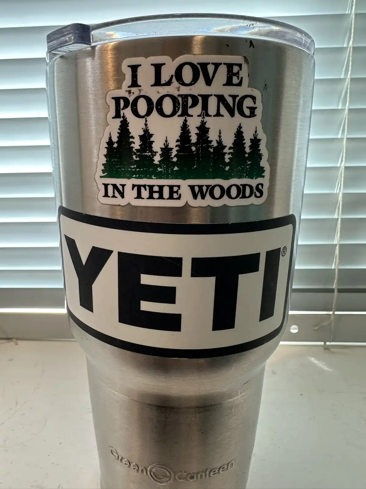 A silver Yeti tumbler with a sticker that reads 'I LOVE POOPING IN THE WOODS' above a graphic of pine trees, placed in front…