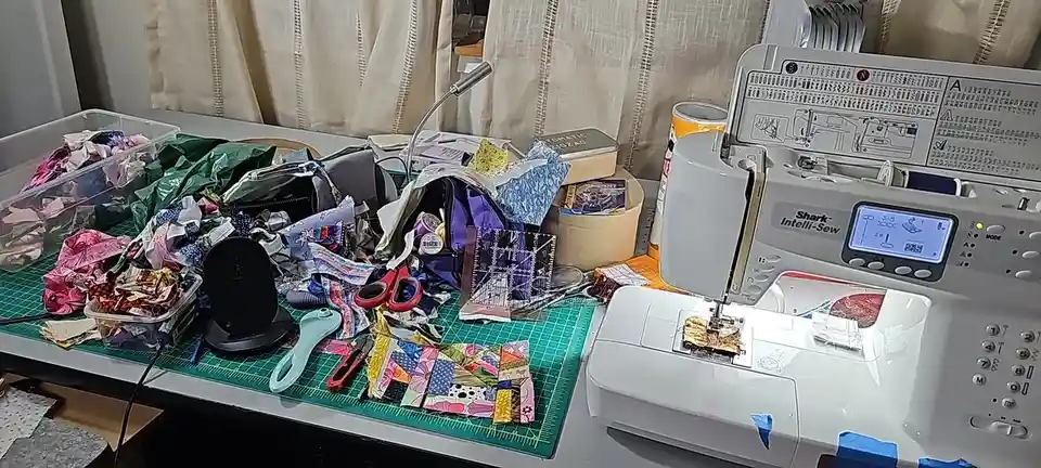 A cluttered sewing workspace features a white Shark Intelli-Sew sewing machine on the right, with a green cutting mat…