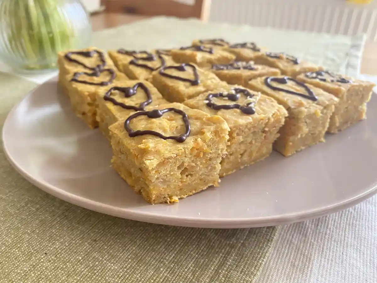 A plate of square, golden-brown baked goods, likely carrot cake bars, each decorated with a dark chocolate heart. The bars…