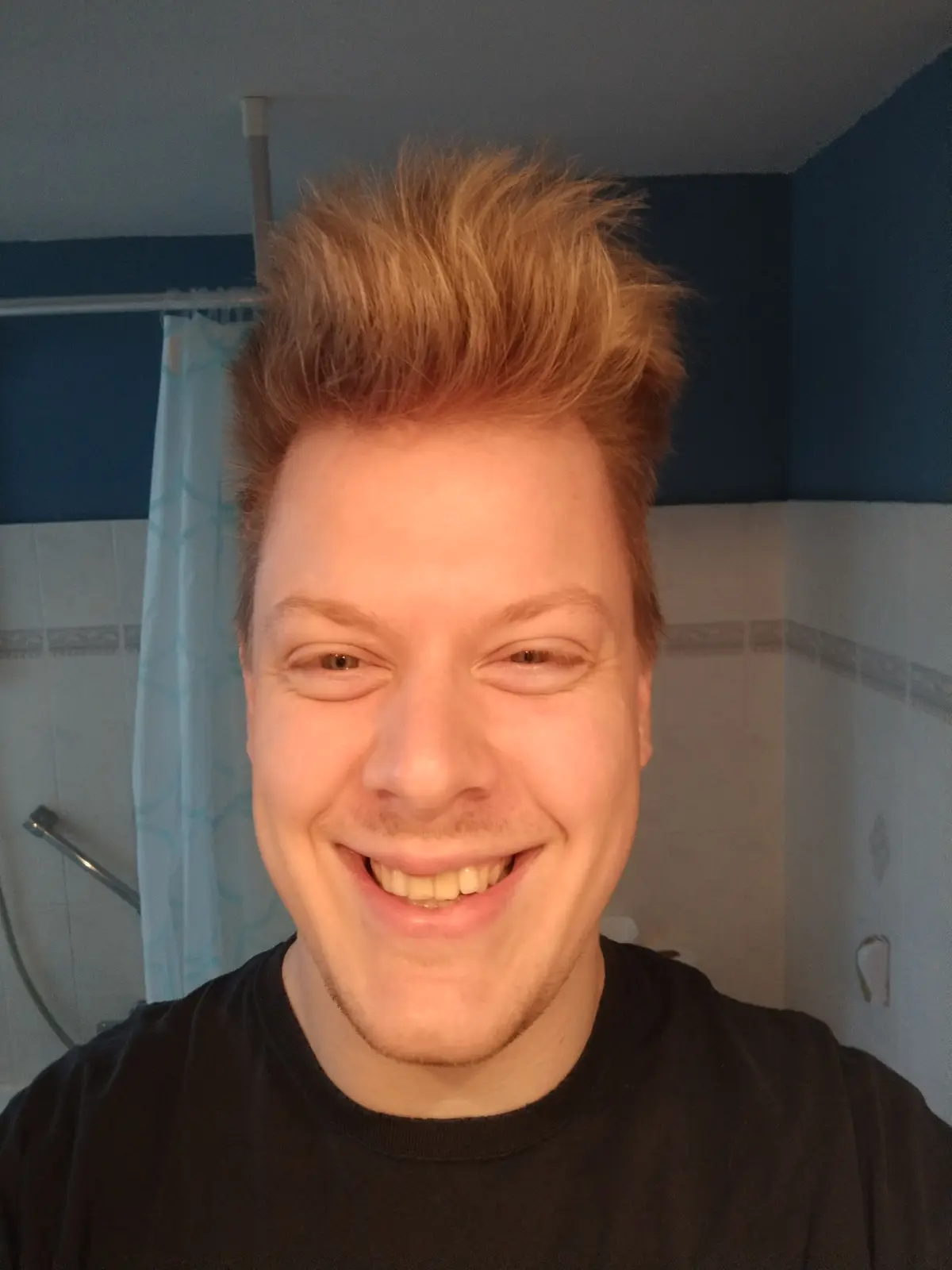 A selfie of a smiling young man with spiky reddish-brown hair in a bathroom with blue walls and a shower curtain. He wears a…