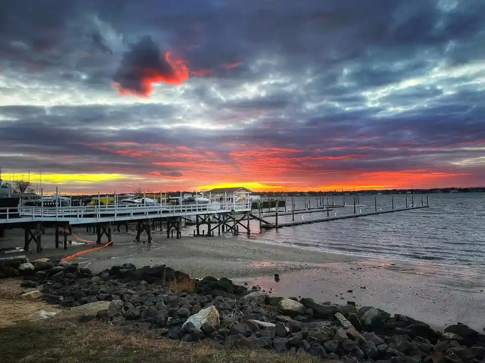 A scenic photo of a dock at sunset with vibrant red and orange clouds over water, featuring a white pier, boats, and rocks…