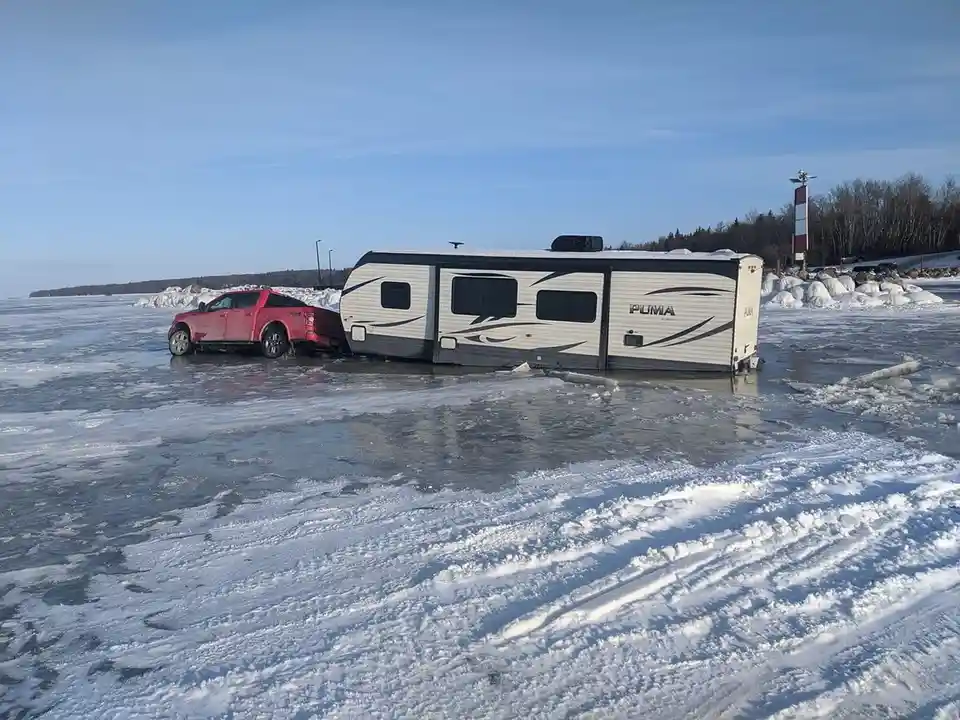 A red pickup truck is stuck in a frozen lake, pulling a white and black 'PUMA' brand RV that is also partially submerged in…