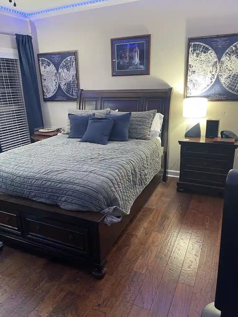 A bedroom with dark wood furniture, blue and grey bedding, and walls decorated with framed vintage-style world maps and a…