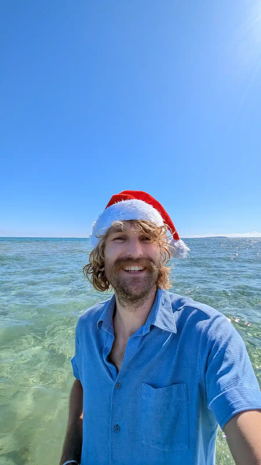 A smiling man with curly hair and a beard wears a red and white Santa hat while taking a selfie in clear, shallow ocean…