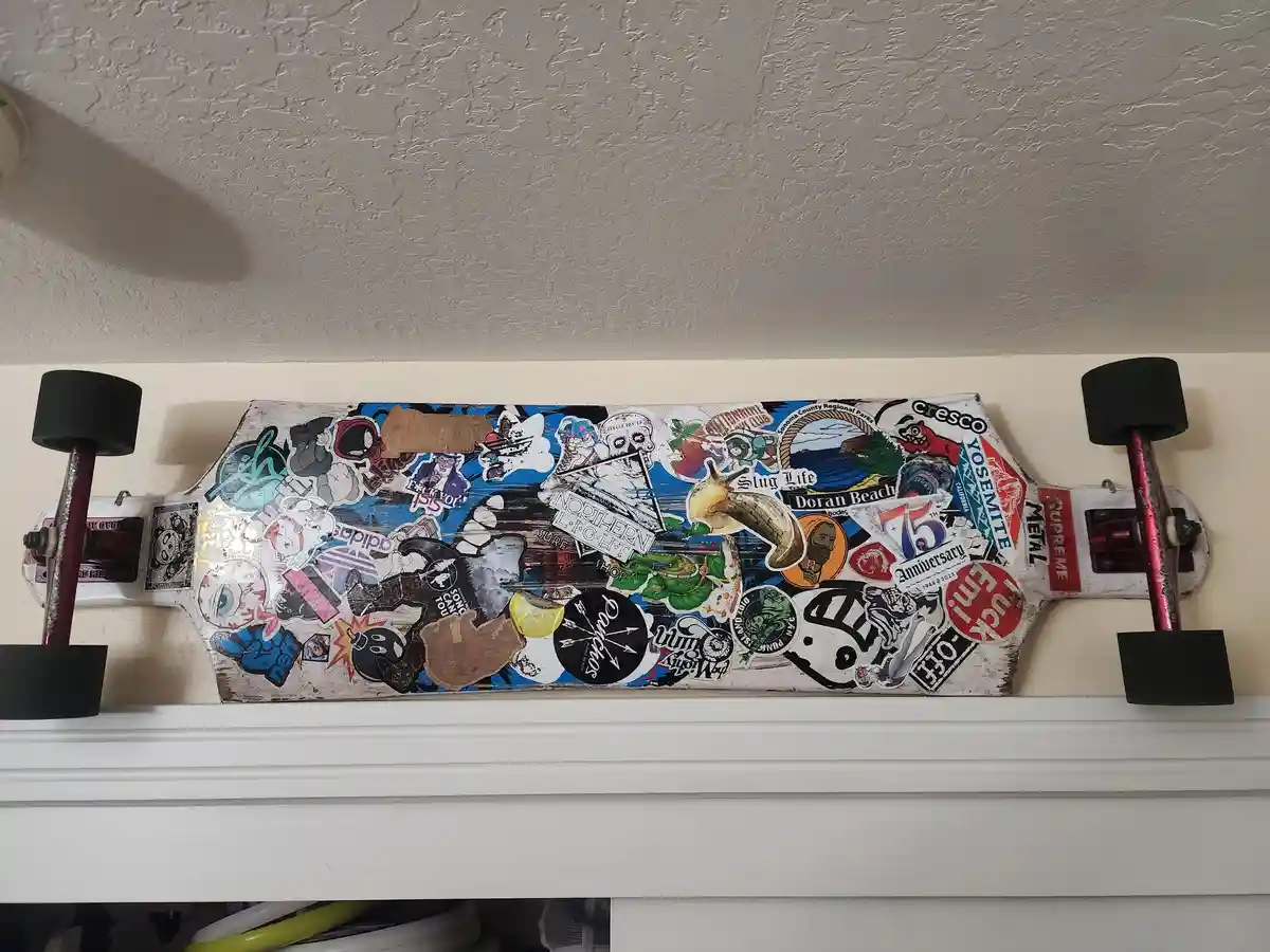 A longboard skateboard, heavily covered in a dense collage of colorful stickers from brands like Supreme and Neff, locations…