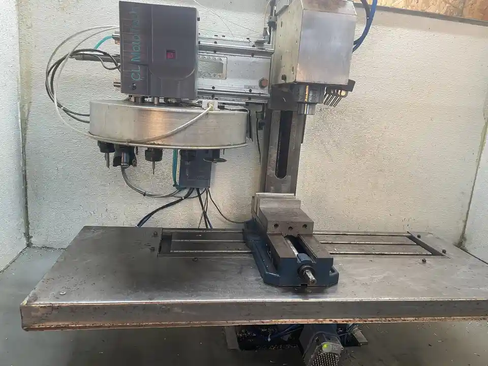 A photo of a used CNC milling machine with a tool changer, showing a workholding vise on its table and various wires and…
