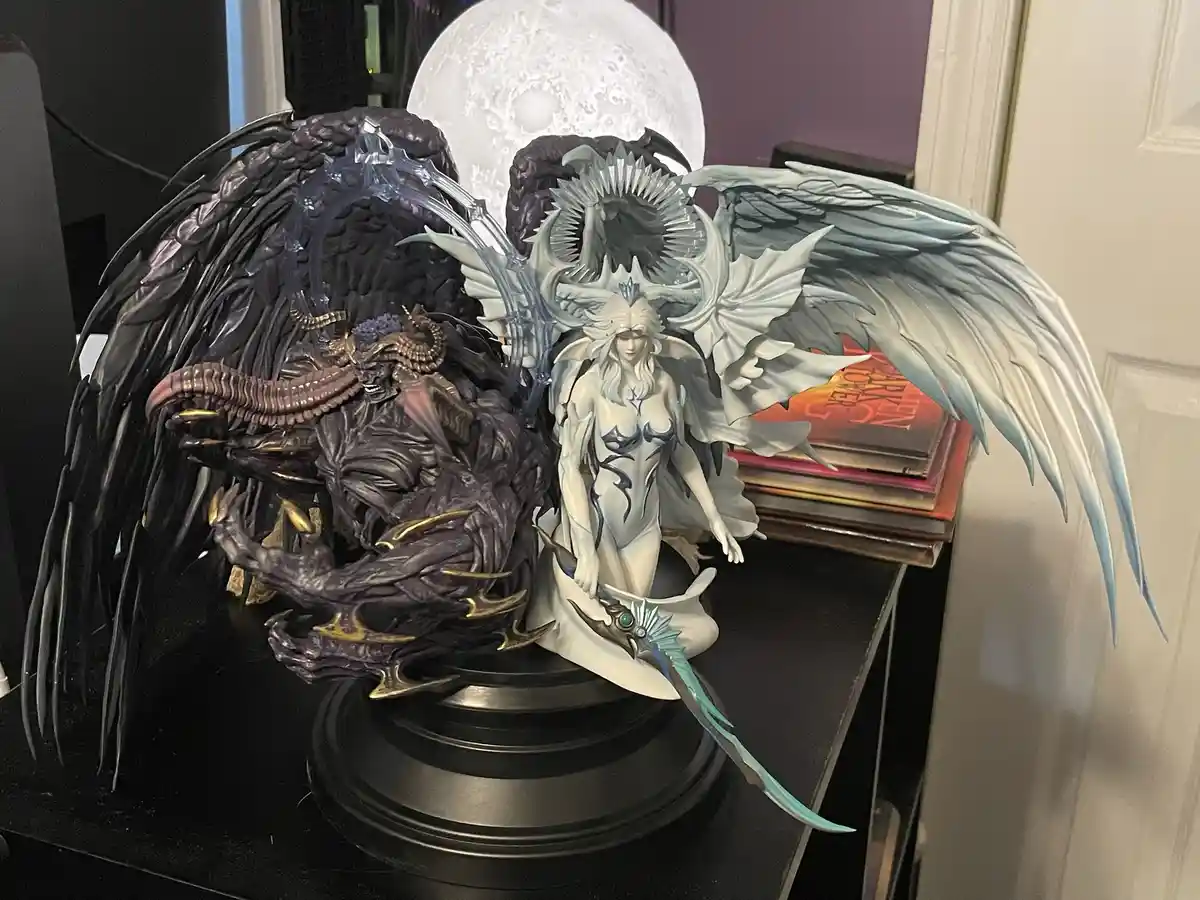 A detailed figurine of a demonic entity with dark wings and horns stands beside a white angelic figure with large, feathered…