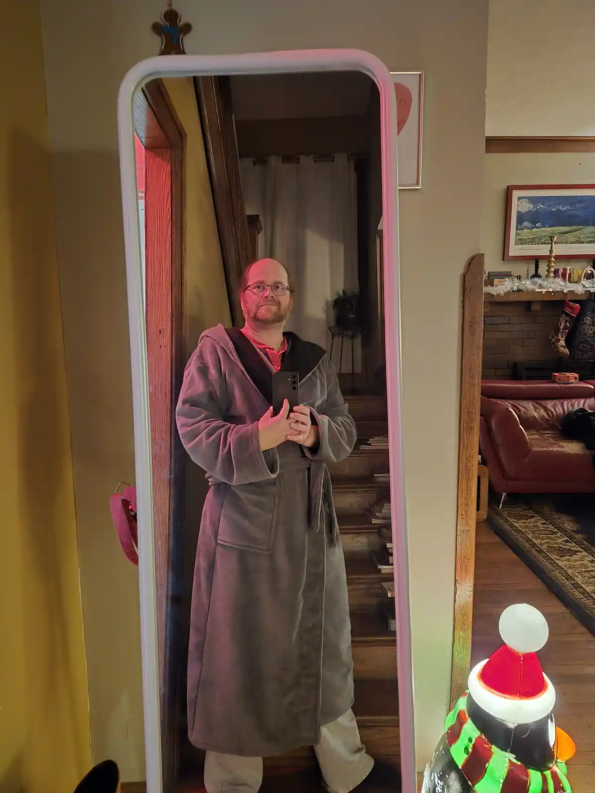 A man in a grey bathrobe and glasses takes a mirror selfie in a home with Christmas decorations, including a lit snowman and…